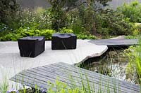 View of terrace made from sliced boulders with abstracted paving design, wooden seats and bridge next to the pond surrounded by Silene latifolia -white campio, Onoclea sensibilis -sensitive fern AGM and Pinus banksiana - jack pine - The Royal Bank of Canada Garden - RHS Chelsea Flower Show 2017 - Designer:  Charlotte Harris - Sponsor: RBC