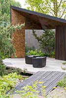 Bridge formed from angular granite blocks leading to a burnt Larch Loggia with cubed seating - The Royal Bank of Canada Garden - RHS Chelsea Flower Show 2017 - Designer: Charlotte Harris - Sponsor: RBC