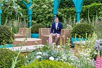 Designer Lee Bestall sitting on one of the wooden slatted benches on the garden with softly clipped yew balls  and roses featured in the surrounding planting. Cornus kousa behind - The Sir Simon Milton Foundation Garden: '500 years of Covent Garden' - RHS Chelsea Flower Show 2017 - Designer: Lee Bestall - Sponsor: Capco Covent Garden