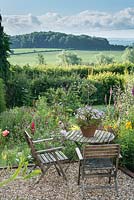 Wooden table and chairs in country garden with view across to open countryside. Viola in terracotta pot.