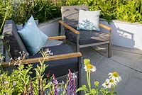 Contemporary sunken patio with seating area surrounded by raised beds with grey armchairs and blue cushions - The Urban Rain garden. RHS Hampton Court Palace Flower Show 2017. Design: Rhiannon Williams. Sponsors: Landform