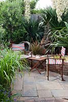 Striped director chairs and small table on paved patio, with Yucca gloriosa flowering in late summer. Bronze Phormium, grasses, fishing net and rope decorations