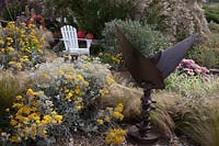 Seaside themed front garden with white Adirondack chair, planted with coastal plants, Cineraria 'Silver Dust', Stipa tenuissima, Erigeron 'Pink Jewel' and Tamarisk tetandra flowering behind. Decorated with driftwood sculptures,  lobster pots, fishing floats and an old rusted propeller