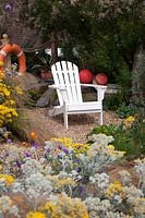 Seaside themed front garden with white Adirondack chair, planted with coastal plants,  including Cineraria 'Silver Dust' , Stipa tenuissima and Tamarisk tetandra flowering behind  and decorated with  life ring and fishing floats. 