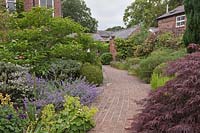 Red brick curving pathway with shrubs and perennials and buildings beyond - June, Cheshire