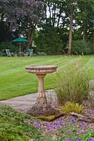 Traditional pedestal stone bird bath in front of lawn with steamer chairs and parasol - June, Cheshire