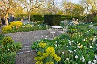 Patio surrounded with spring borders of tulips, daffodils, Mediterranean spurge, lunaria - honesty, forget me nots and hellebores. Thea Maldegem garden
