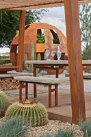 A view through a corroded steel frame pergola and table and benches made with battered edge sandstone tops to rusted steel dome with Echinocactus grusonii and other drought-tolerant cacti and perennials in the Cactus Direct:2101 Garden at Tatton RHS Flower Show 2017