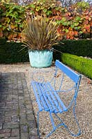 A bright blue painted bench sits on pea shingle and brick  backed with Vitis coignetiae, Taxus baccata - Yew - hedge and a lower Buxus sempervirens  - Box - hedge. Phormium tenax purpureum group planted in old copper tub.