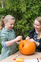 A young girl removes the cutout section of an eye on a large pumpkin, helped by her mother