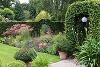 A hornbeam arch straddles a path leading to an arch cut in a long hedge, partly hidden behind a bed of pink and blue perennials.