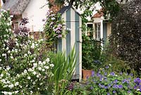 A wooden tool shed is painted in stripes, and Clematis 'Hagley Hybrid' trained up the left side. In front stands fragrant philadelphus.