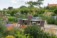 Stone terrace with dining table is edged in orange Geum, Catmint, Cirsium, hardy Geranium, Valerian, Salvia and Stipa gigantea. Beyond, beech hedge containing Cherry Walk.