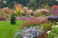 Garden view of flower beds with Aster x frikartii 'Monch', Persicaria amplexicaulis 'Atrosanguinea'. Plus chairs facing bed of grasses, wooden gazebo 