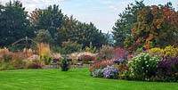 Autumn beds and borders full of flowering perennials and grasses against a backdrop of deciduous trees with lawn in foreground
