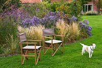Lawn with seats and pet dog, with perennial border of grass Stipa tenuissima, Aster 'Little Carlow' and Verbena bonariensis.