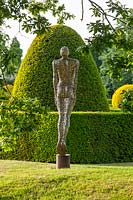 Sculpture of a figure in stainless steel on a lawn with topiary backdrop
