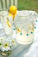 Jug of lemonade with crocheted beaded jug cover, drink tumblers, lemons and small vase of cut flowers