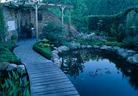 Pond with Nymphaea ( water lilies ), Pontederia ( Hecht herb ), edged with natural stone, Boardwalk as a bridge, bedding with Primula vialii ( Orchid primroses ), Buxus ( book ) as a mini-hedge, seating on patio with brick wall
