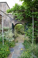 Charingworth Court, Glos, summer, paved path leading trhough arched gate beneath bamboo pergola