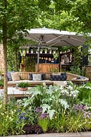 The Gaze Burvil Garden. A modular sofa with cushions sits beneath a large sunshade. Beyond lies an outdoor kitchen with barbecue, pans and pots of herbs. Designer: Ross Allan. Sponsor: Gaze Burvill.