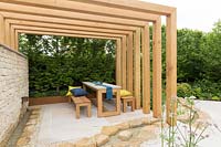 The Kampo No Niwa Garden, view of wood pergola with wooden table and bench seats and cushions, stone patio with water feature rill, dry stone wall with waterfall – Designer: Kazuto Kashiwakura and Miko Sato- Sponsor: Kampo No Niwa 300 sponsors. RHS Chelsea Flower Show 2019.