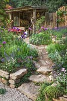 Stone steps and path leading to a wooden lean to shelter in a Mediterranean style planted garden. Stone walled raised beds with purple and pink flowering plants and silver foliage plants. The Donkey Sanctuary: Donkeys Matter Design: Christina Williams and Annie Prebensen. Sponsor: The Donkey Sanctuary. RHS Chelsea Flower Show 2019