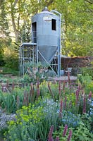 The Resilience Garden at RHS Chelsea Flower Show 2019. View of the garden with the repurposed grain silo and planting which includes Linum perenne, Echium russicum and Euphorbia seguierina subsp Niciciana - Designer: Sarah Eberle - Sponsors: Gravetye Manor Hotel and Restautant, Kingscote Estate, Forestry Commission, Royal Botanic Gardens, Kew, Scottish Foresty, Welsh Governement