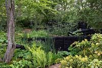 The M and G Garden. A lush woodland with blackened timber sculptures by Johnny Woodford, creating a dramatic backdrop for green understorey planting. Awarded an RHS Gold Medal. Designer: Andy Sturgeon. Sponsor: M and G - Chelsea Flower Show 2019