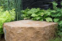 The M and G Garden, view of green planted woodland garden with shade loving plants English ironstone stone seat, charred oak walls sculpture by Johnny Woodford – Designer: Andy Sturgeon - Sponsor: M and G 