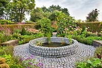 Circular raised pond made of stone setts with marginal planting and formal mixed borders.