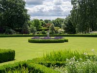A view across garden lawn, with stone urn focal point, surrounded by a circular hedge of Buxus sempervirens and flowering Lavandula. 

