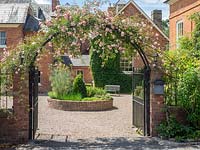 An iron arch covered with climbing pink Rosa, through which a raised herb bed can be seen.
