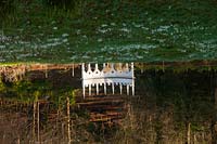 A reflection in lake of an exedra, naturalised Galanthus and woodland beyond. 