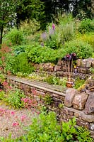 A recessed wooden bench set into a stone wall surrounded by self seeded Erigeron karvinskianus, campanula, sedum and Centranthus ruber, Church View, Appleby-in-Westmorland, Cumbria, UK.