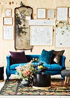 A wall of framed illustrations behind a blue sofa, table and fresh flowers. 