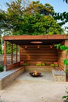  A contemporary pergola with fire pit and wooden bench seating. 
