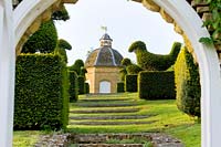 View through arch to set of grassed steps bordered by clipped topiary birds, leading up to stone dovecot.