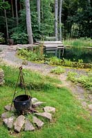 View of handmade fire pit nearby a natural swimming pool surrounded by tall forest trees.