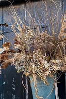 Selection of sprayed gold dried seedheads, ferns, berries and leaves displayed in florist's bucket hanging on black door