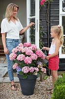 Girl and woman enjoying each other's company on patio whilst girl waters hydrangea in a pot
