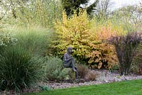 Statue of a seated boy set on boulder in gravel bed with backdrop of Cornus - dogwood. Other plants as specimens in gravel include: Berberis Miscanthus