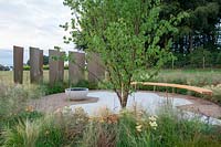 Prunus serrula in morning light, gold coloured glass screens surround a circular gravel and paved patio with curved bench, planting of Astrantia major' Claret', Achillea 'Terracotta', Molinia caerulea subsp. caerulea 'Heidebraut' and Deschampsia cespitosa 'Goldtau' - JW Lees: From Hop to Glass Garden, RHS Tatton Park Flower Show 2018