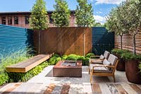 Contemporary seating area with fireplace surrounded by Olea europaea and
 Muehlenbeckia complexa in rusted steel containers, Quercus ilex, Buxus 
sempervirens balls topiary by rusted panel wall and Miscanthus sinensis
 'Morning Light' by the bench. 