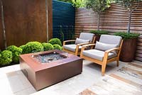 Contemporary seating area with fireplace, chairs next to big rusted corten 
steel containers with Olea europaea and Muehlenbeckia complexa and Buxus 
sempervirens balls topiary by wooden and rusted panel walls. 