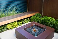 Contemporary seating area with fireplace, and wooden bench by blue wall and 
rusted panels surrounded by Miscanthus sinensis 'Morning Light', Dryopteris 
erythrosoraand Buxus sempervirens balls topiary. 