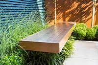 Wooden bench surrounded by Miscanthus sinensis 'Morning Light', Dryopteris 
erythrosora and Buxus sempervirens balls topiary by rusted panel wall. 