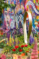 Metal ribbon sculpture against colourful grafitti wall and mixed planting. The Supershoes, Laced with Hope Garden, a partnership with Frosts. Sponsor: Frosts Garden Centres, RHS Chelsea Flower Show, 2018.