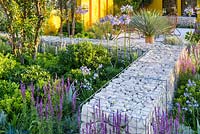 Stone-filled gabion wall with multi-stemmed Arbutus unedo. 'Living La Vida 120'. RHS Hampton Flower Show 2018