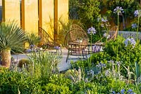 Chairs on stone terrace with yellow wall, Agapanthus 'Blue Storm' and Stachys byzantina. 'Living La Vida 120'. RHS Hampton Flower Show 2018 